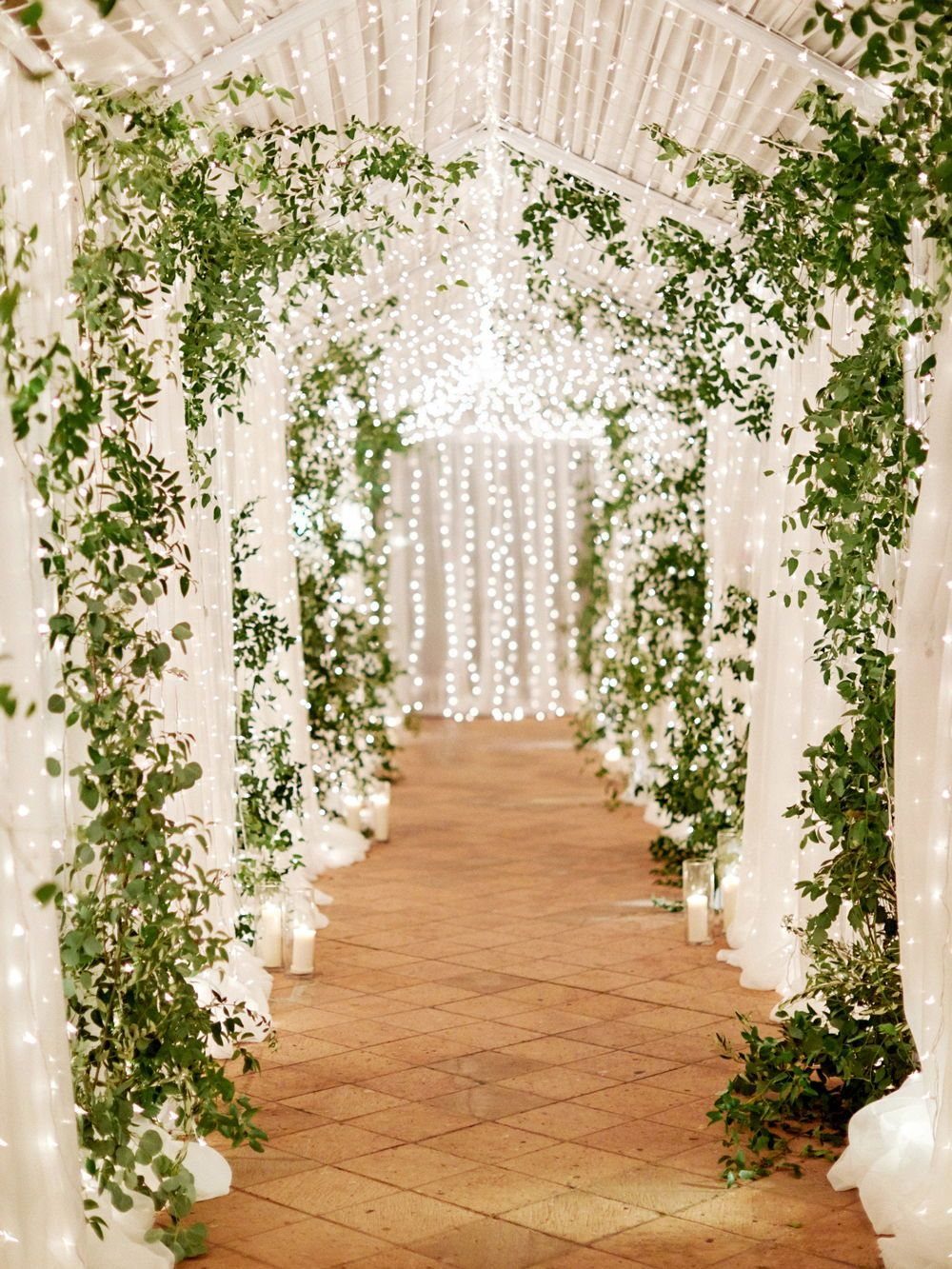 Light tunnel with white draped fabric, warm fairy lights, and green leafy vines.
