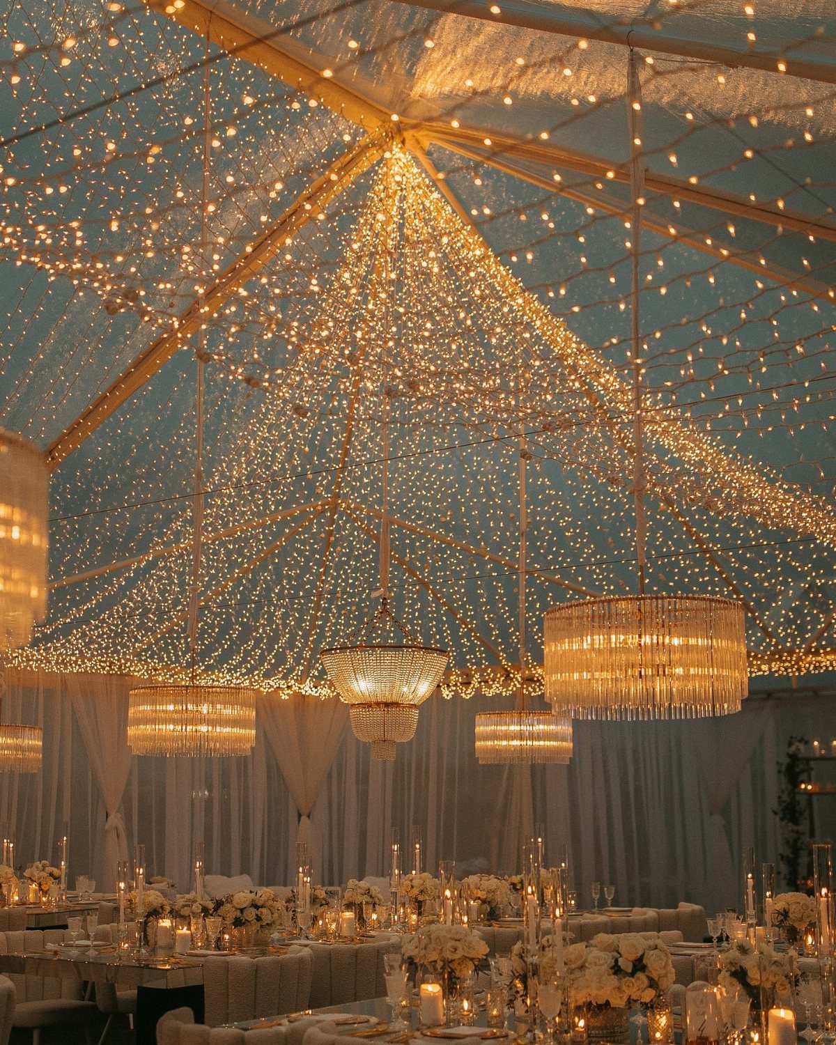 Wedding tent ceiling covered in warm white fairy lights and crystal chandeliers.