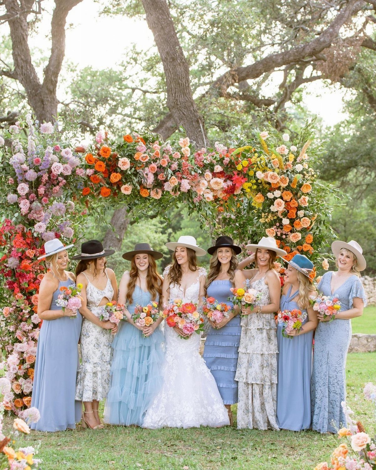 Bride and bridesmaids in western attire wearing cowboy hats under a floral arch.