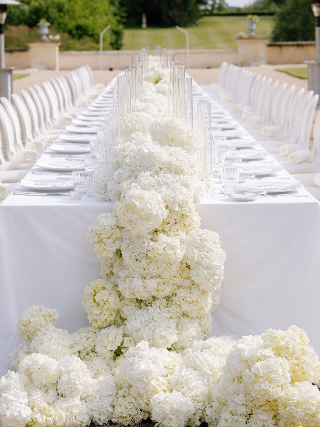 White hydrangea flower runner cascading down a long white wedding banquet table outdoors.