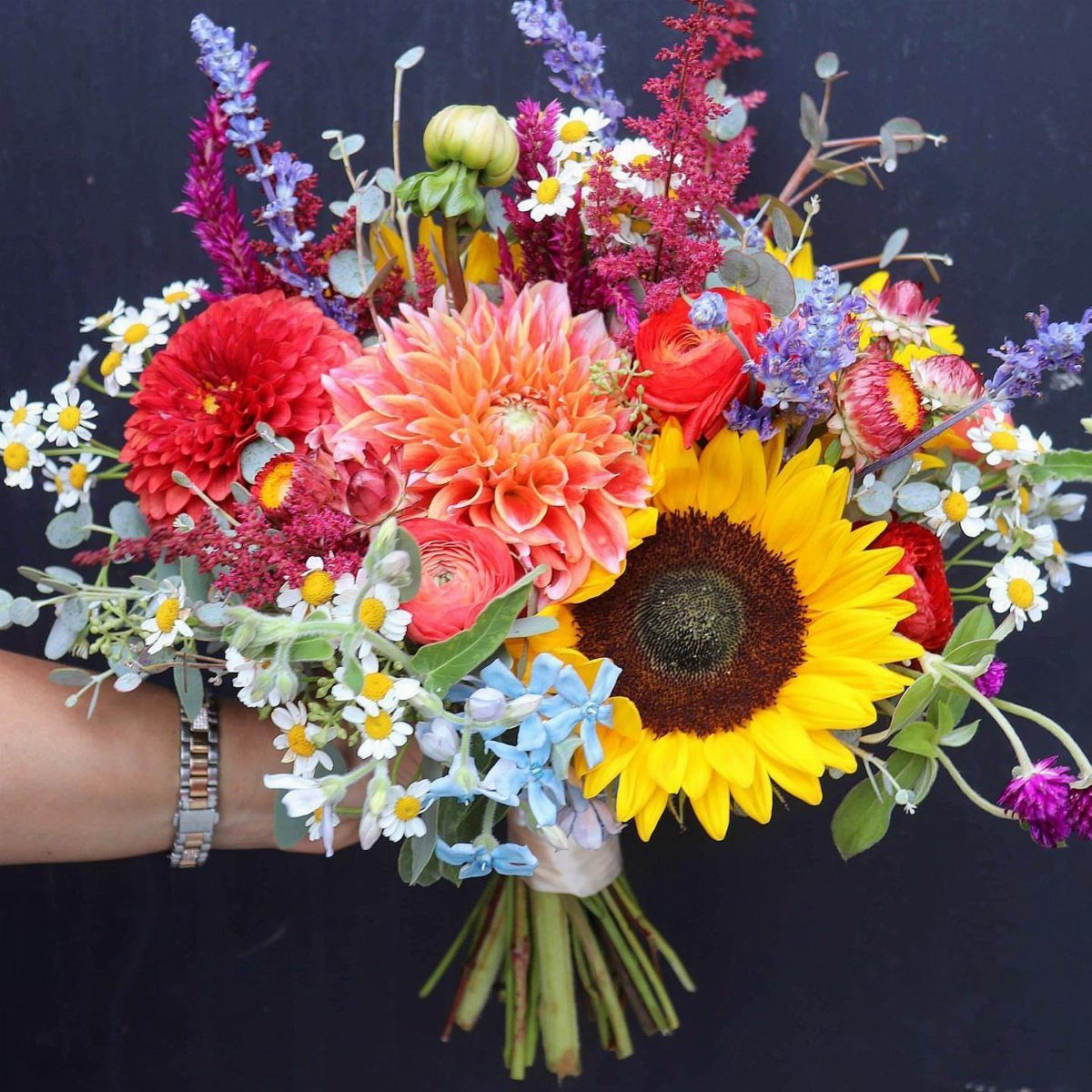 Colorful bridal bouquet with sunflowers, orange dahlias, and purple wildflowers against dark background.