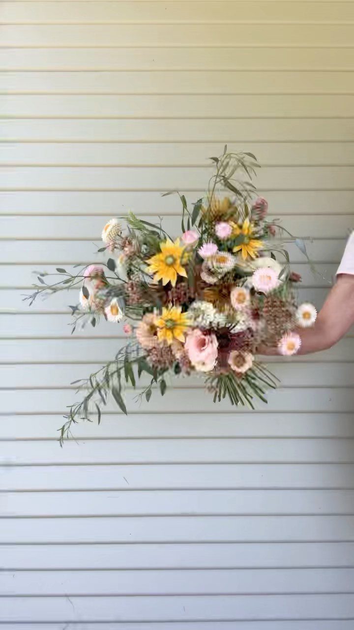 Wildflower bridal bouquet with sunflowers, pink roses, and white daisies against white siding.