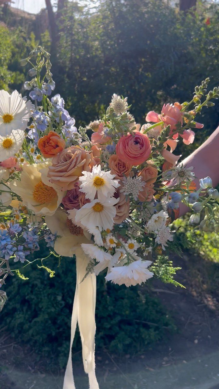 Bridal bouquet with white daisies, peach roses, and blue delphinium tied with ribbon.