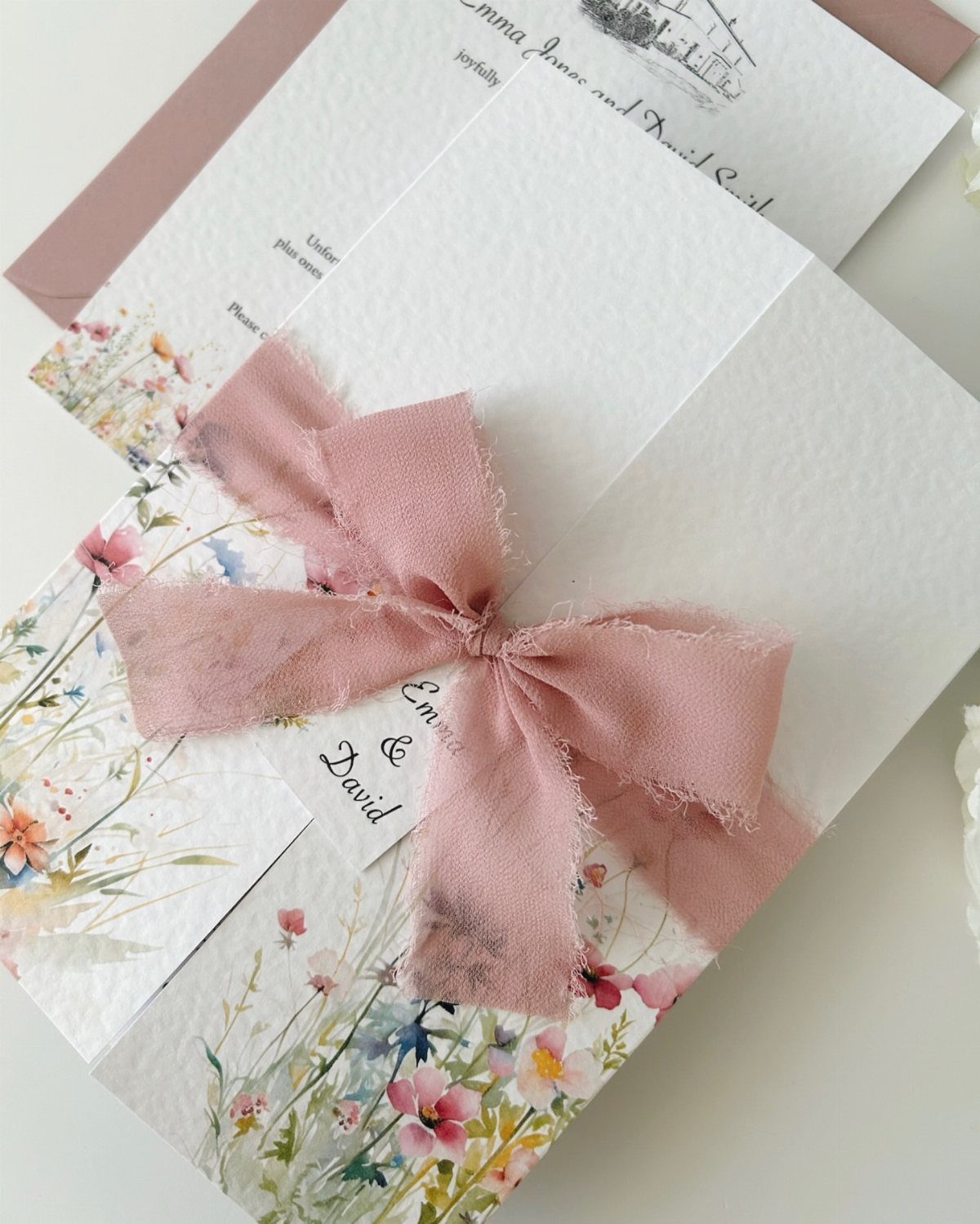 Wedding invitations with wildflower illustrations tied with a dusty pink ribbon bow.