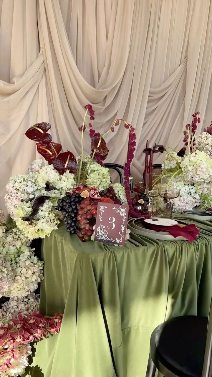 Wedding table with green cloth, burgundy and white flowers, and cream draped backdrop.